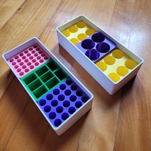 May include: Two rectangular, white storage containers with colorful inserts. One has pink, green, and purple compartments. The other has yellow and purple sections. The containers are on a wooden surface.