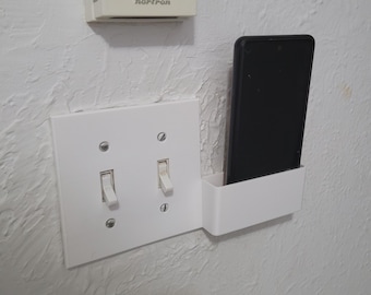 May include: A white light switch plate with two toggle switches and a white phone holder attached to the right side. A black smartphone is in the holder.