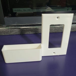 May include: White plastic wall mount with a rectangular opening and a smaller rectangular shelf below. The mount has two small holes on the top and bottom.