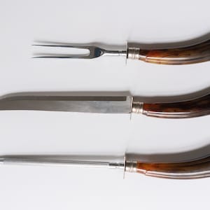 May include: A carving set featuring a fork, a knife, and a sharpening steel. The stainless steel blades and tines are paired with handles of dark brown wood. Each handle is accented with a silver-colored bolster.
