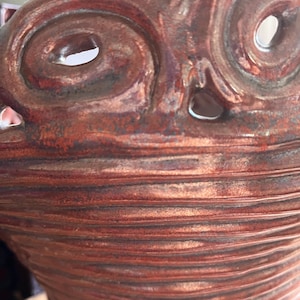 Op de afbeelding: Close-up van een handgemaakte, roodbruine keramische pot met een uniek, getextureerd ontwerp. De pot heeft een gezichtachtige vorm met oogachtige openingen en een geribbelde, getextureerde body. Het keramiek heeft een glanzende afwerking.