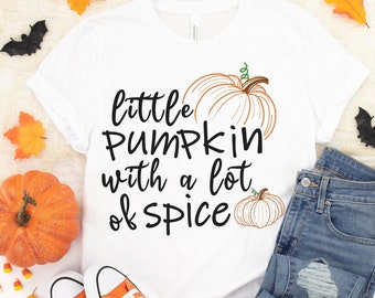 Puede incluir: Camiseta blanca con el texto "little pumpkin with a lot of spice" y dos ilustraciones de calabazas naranjas. La camiseta está colocada sobre una superficie blanca con un par de zapatillas naranjas, unos vaqueros azules, una calavera y hojas de otoño.