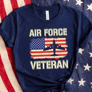 Pode incluir: Camiseta azul marinho com o texto "AIR FORCE VETERAN", um desenho da bandeira americana e uma silhueta de avião. A camiseta é mostrada em um fundo da bandeira americana.