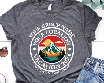 May include: A gray t-shirt with a white circle design featuring a mountain scene with a sunset, the text "Your Group Name Your Location Vacation 2024 Mountain | Lake | Camp" and a white border.