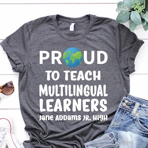 May include: A dark gray t-shirt with white text that reads "Proud to teach multilingual learners Jane Addams Jr. High". The shirt features a small graphic of a blue and green globe.