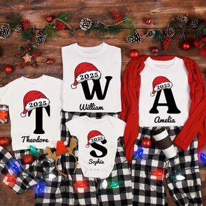 Pode incluir: Uma coleção de camisetas e pijamas de Natal brancos com calças xadrez em preto e branco. Cada camisa apresenta uma grande inicial, um gorro de Papai Noel com "2025" e um nome. Decorações festivas de feriados cercam as roupas.
