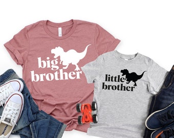 May include: Two t-shirts with dinosaur graphics and text. The larger shirt is pink with white text that says "big brother" and a white dinosaur. The smaller shirt is gray with black text that says "little brother" and a black dinosaur.