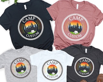 May include: Assorted t-shirts in navy, dusty rose, gray, white, and charcoal. Each shirt features a circular graphic with the words "CAMP CUSTOM" around a sunset scene with trees and a tent. The shirts are short-sleeved.