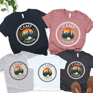 Pode incluir: V&aacute;rias camisetas de manga curta em azul marinho, rosa empoeirado, cinza, branco e carv&atilde;o. Cada camisa apresenta um gr&aacute;fico circular com as palavras "CAMP CUSTOM" em torno de uma cena de p&ocirc;r do sol com &aacute;rvores e uma tenda.