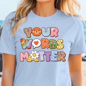 Pode incluir: T-shirt azul claro com a frase "YOUR WORDS MATTER" em letras coloridas e divertidas. O design inclui um urso, um coração, flores e listras de arco-íris. A camisa é feita de um material macio.