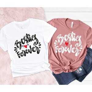 Pode incluir: Duas camisetas com o texto "Besties Forever" e um coração vermelho. As camisetas são brancas e rosadas.