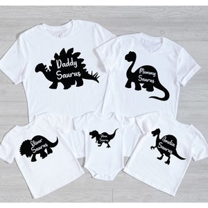 May include: White t-shirts and a onesie with black dinosaur silhouettes and the names "Daddy Saurus", "Mommy Saurus", "Oliver Saurus", "Olivia Saurus", and "Amelia Saurus". The shirts have short sleeves and a crew neck.