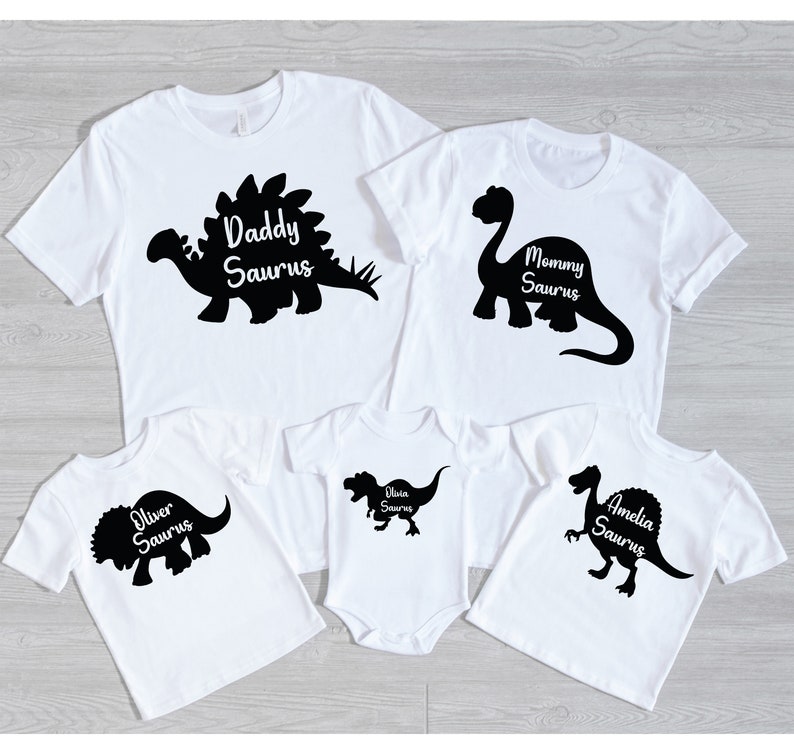May include: White t-shirts and a onesie with black dinosaur silhouettes and the names "Daddy Saurus", "Mommy Saurus", "Oliver Saurus", "Olivia Saurus", and "Amelia Saurus". The shirts have short sleeves and a crew neck.