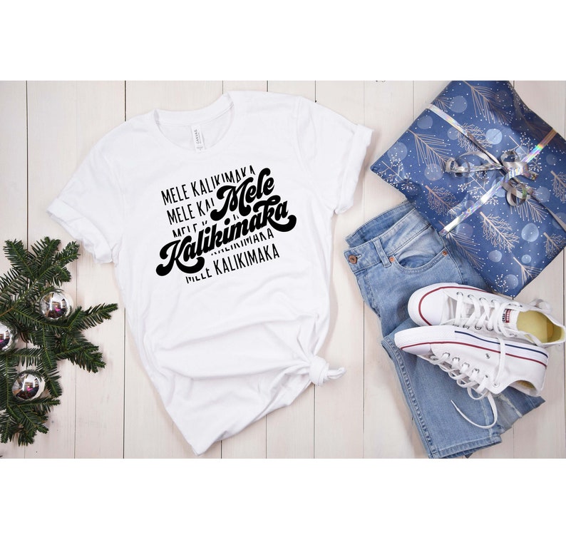May include: White t-shirt with the phrase "Mele Kalikimaka" in black script. The shirt is styled with blue jeans, white sneakers, and a wrapped gift. A Christmas tree branch with silver ornaments is in the background.