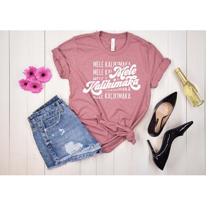 May include: A dusty rose t-shirt with the text "Mele Kalikimaka" in white script, paired with denim shorts, black high heels, pink flowers, and a bottle of wine. The items are arranged on a white wooden surface.