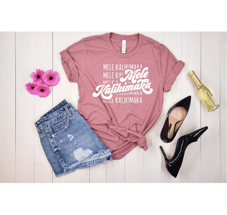 May include: A dusty rose t-shirt with the text "Mele Kalikimaka" in white script, paired with denim shorts, black high heels, pink flowers, and a bottle of wine. The items are arranged on a white wooden surface.