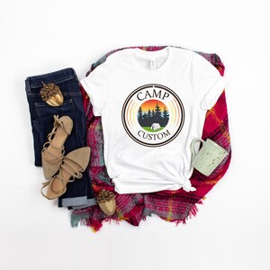 Pode incluir: Camiseta branca com um gr&aacute;fico circular com tema de acampamento, apresentando &aacute;rvores, uma tenda e um p&ocirc;r do sol, com o texto "CAMP CUSTOM". A camiseta &eacute; exibida com jeans azuis, sand&aacute;lias bege e um cachecol xadrez vermelho.