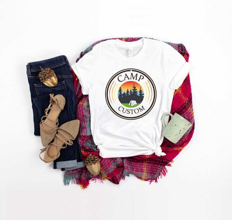 Pode incluir: Camiseta branca com um gr&aacute;fico circular com tema de acampamento, apresentando &aacute;rvores, uma tenda e um p&ocirc;r do sol, com o texto "CAMP CUSTOM". A camiseta &eacute; exibida com jeans azuis, sand&aacute;lias bege e um cachecol xadrez vermelho.