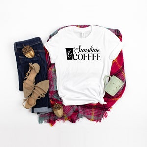 May include: White t-shirt with the text "Sunshine & COFFEE" and a coffee cup graphic. The shirt is laid out on a red and white plaid blanket with a pair of brown sandals, blue jeans, and a small green mug.