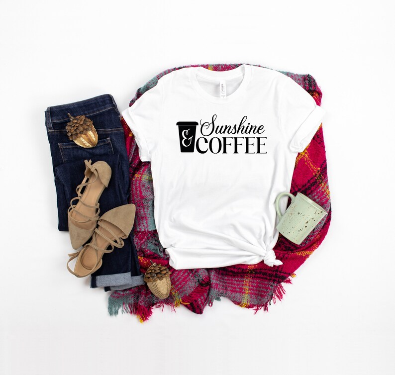 May include: White t-shirt with the text "Sunshine & COFFEE" and a coffee cup graphic. The shirt is laid out on a red and white plaid blanket with a pair of brown sandals, blue jeans, and a small green mug.