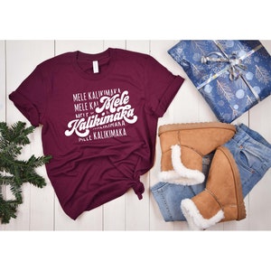 May include: A maroon t-shirt with the words "Mele Kalikimaka" in white script. The shirt is paired with light brown boots with white faux fur trim, blue jeans, and a wrapped gift with a blue and silver design.