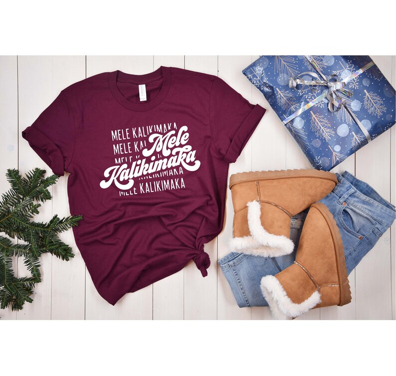 May include: A maroon t-shirt with the words "Mele Kalikimaka" in white script. The shirt is paired with light brown boots with white faux fur trim, blue jeans, and a wrapped gift with a blue and silver design.