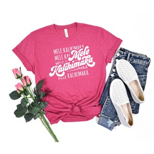 May include: A magenta t-shirt with the text "Mele Kalikimaka" in white script. The shirt is styled with a pair of blue jeans, white slip-on shoes, and a stem of pink roses. The shirt is knotted at the waist.