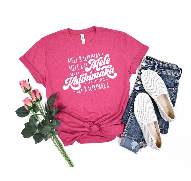 May include: A magenta t-shirt with the text "Mele Kalikimaka" in white script. The shirt is styled with a pair of blue jeans, white slip-on shoes, and a stem of pink roses. The shirt is knotted at the waist.