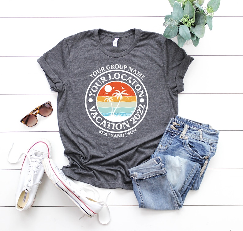 May include: A dark gray t-shirt with a white circle graphic featuring palm trees, a sunset, and the text "Your Group Name Your Location Vacation 2022 Sea | Sand | Sun".