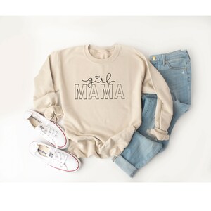 Peut inclure: Un sweat-shirt beige avec le texte "girl mama" en lettres noires. Le sweat-shirt est pos&eacute; sur une surface blanche avec une paire de baskets blanches et un jean bleu.