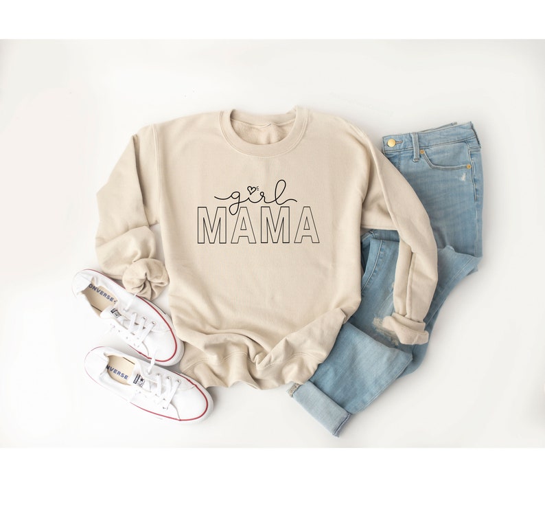Peut inclure: Un sweat-shirt beige avec le texte "girl mama" en lettres noires. Le sweat-shirt est pos&eacute; sur une surface blanche avec une paire de baskets blanches et un jean bleu.