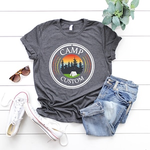 Pode incluir: T-shirt cinzento escuro com um gr&aacute;fico circular com um p&ocirc;r do sol, &aacute;rvores e uma tenda. As palavras "CAMP CUSTOM" est&atilde;o impressas. A camisa &eacute; combinada com jeans azuis, t&ecirc;nis brancos e &oacute;culos de sol.