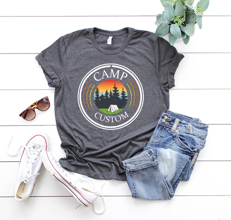Pode incluir: T-shirt cinzento escuro com um gr&aacute;fico circular com um p&ocirc;r do sol, &aacute;rvores e uma tenda. As palavras "CAMP CUSTOM" est&atilde;o impressas. A camisa &eacute; combinada com jeans azuis, t&ecirc;nis brancos e &oacute;culos de sol.