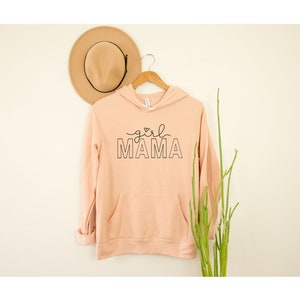 Peut inclure: Un sweat &agrave; capuche couleur p&ecirc;che avec un motif noir qui dit "girl mama" avec un petit c&oelig;ur au-dessus du mot "mama".