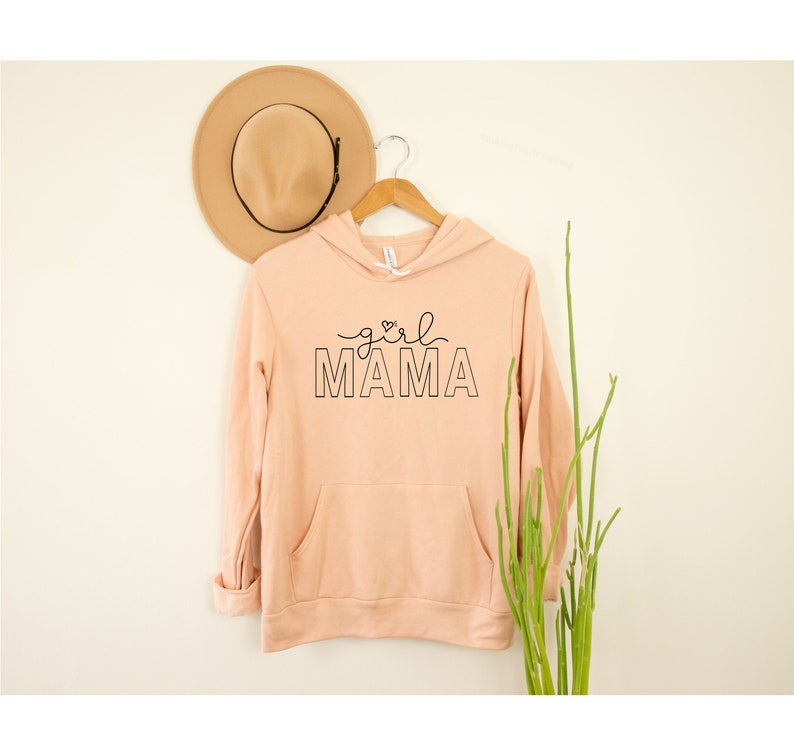 Peut inclure: Un sweat &agrave; capuche couleur p&ecirc;che avec un motif noir qui dit "girl mama" avec un petit c&oelig;ur au-dessus du mot "mama".