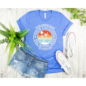 May include: A blue t-shirt with a white circle graphic that says "Your Group Name Your Location Vacation 2022 Sea | Sand | Sun". The graphic features a sunset with palm trees and a beach.