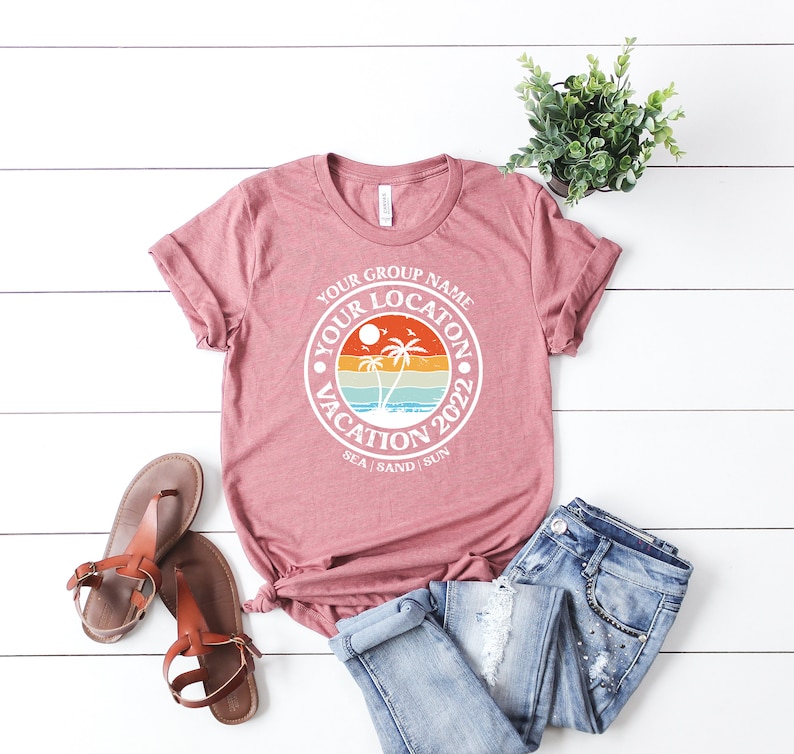 May include: A pink t-shirt with a white circle graphic that says "Your Group Name Your Location Vacation 2022 Sea Sand Sun". The graphic features a sunset with palm trees and a beach.