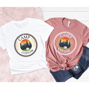 Pode incluir: Duas camisetas, uma branca e outra rosa empoeirado, cada uma com um design gr&aacute;fico circular com as palavras "CAMP CUSTOM". O design inclui um p&ocirc;r do sol, &aacute;rvores e uma tenda, evocando um tema de acampamento. As camisetas s&atilde;o roupas casuais.