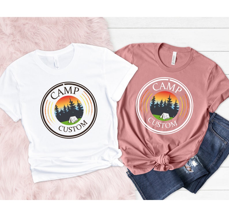 Pode incluir: Duas camisetas, uma branca e outra rosa empoeirado, cada uma com um design gr&aacute;fico circular com as palavras "CAMP CUSTOM". O design inclui um p&ocirc;r do sol, &aacute;rvores e uma tenda, evocando um tema de acampamento. As camisetas s&atilde;o roupas casuais.