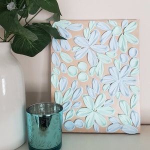 May include: A canvas painting with a textured, raised paint design. The painting features a floral pattern with light blue and mint green flowers on a beige background.