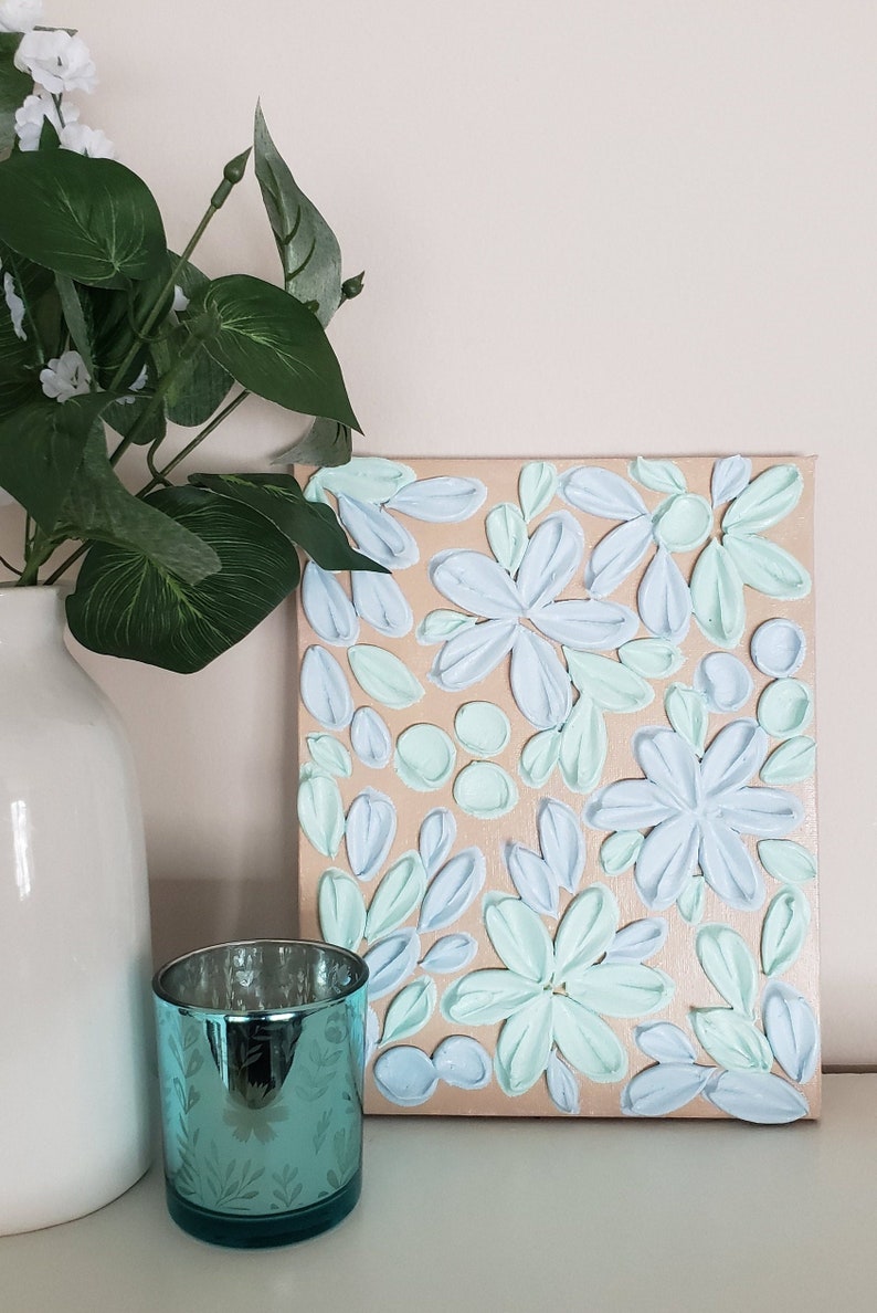 May include: A canvas painting with a textured, raised paint design. The painting features a floral pattern with light blue and mint green flowers on a beige background.