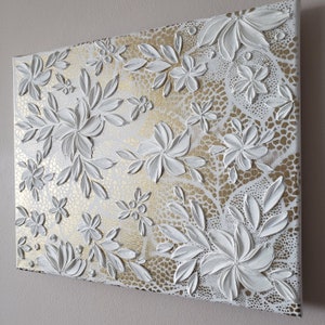 Puede incluir: Pintura floral texturizada blanca sobre fondo dorado. Las flores son en relieve y tienen un efecto 3D.