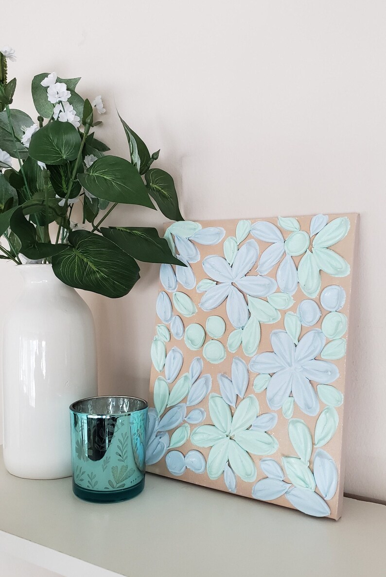 May include: A canvas painting with a textured, raised floral design. The flowers are painted in shades of light blue and green on a beige background.