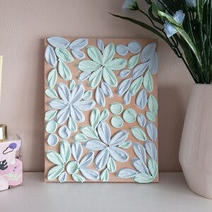 May include: A textured canvas painting with a floral design. The flowers are painted in shades of blue and green on a beige background.