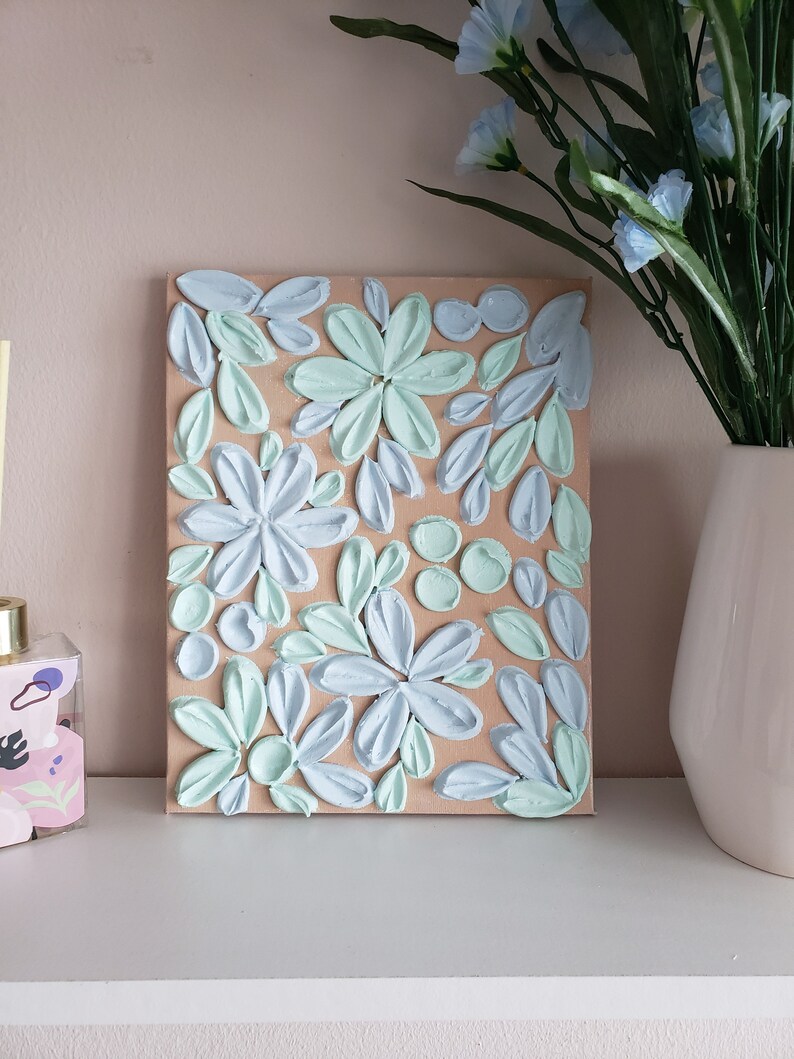 May include: A textured canvas painting with a floral design. The flowers are painted in shades of blue and green on a beige background.