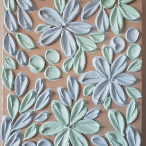 May include: A close-up of a canvas painting with a textured, raised floral design. The flowers are painted in shades of light blue and light green on a beige background.