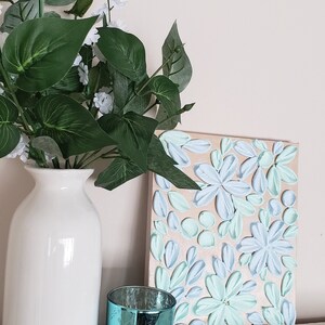May include: A white vase with green leaves and white flowers sits on a white shelf next to a teal candle holder and a canvas painting with a floral design in shades of blue and green.