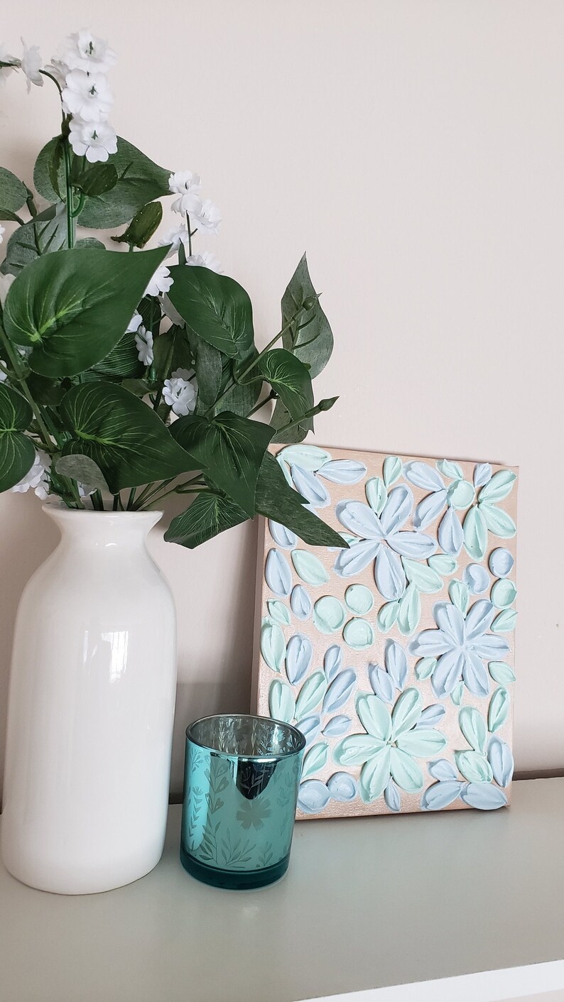 May include: A white vase with green leaves and white flowers sits on a white shelf next to a teal candle holder and a canvas painting with a floral design in shades of blue and green.