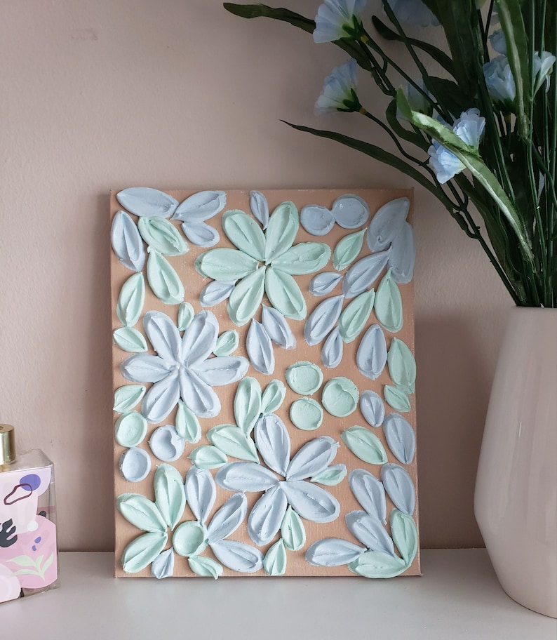 May include: A textured canvas painting with a floral design. The flowers are painted in shades of blue and green, and the background is a light brown color.