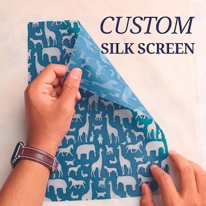 May include: A blue silk screen with a white animal pattern is being held up to reveal the design. The text "CUSTOM SILK SCREEN" is visible in the upper right corner. The design includes giraffes, elephants, and other animals.